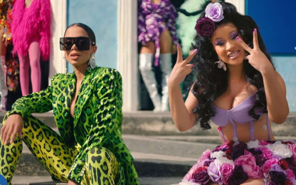 Anitta no lado esquerdo de roupa verde e óculos escuros e Cardi B no lado direito com roupa roxa sentadas em uma escada no clipe da música Me Gusta