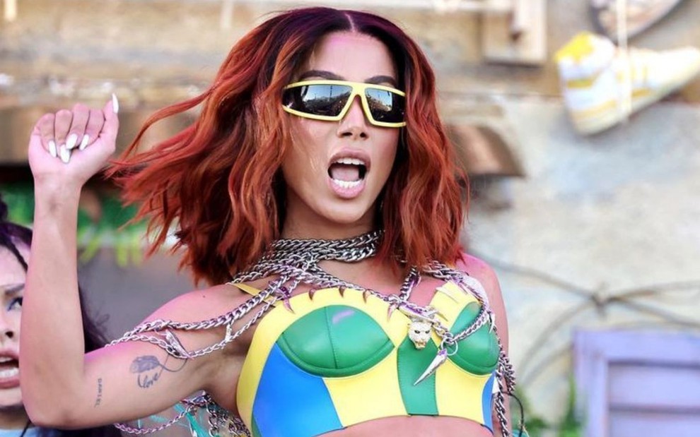 Anitta de cabelos ruivos veste óculos de sol e top com as cores do Brasil
