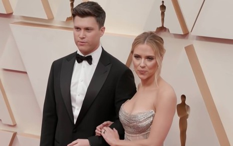 Colin está ao lado de Scarlett, ele usa terno preto e camiseta branca com gravata borboleta; Scarlett olha para o lado e usa vestido prata