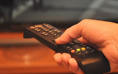 Imagem mostra a mão de uma pessoa segurando um controle remoto apontado para uma televisão