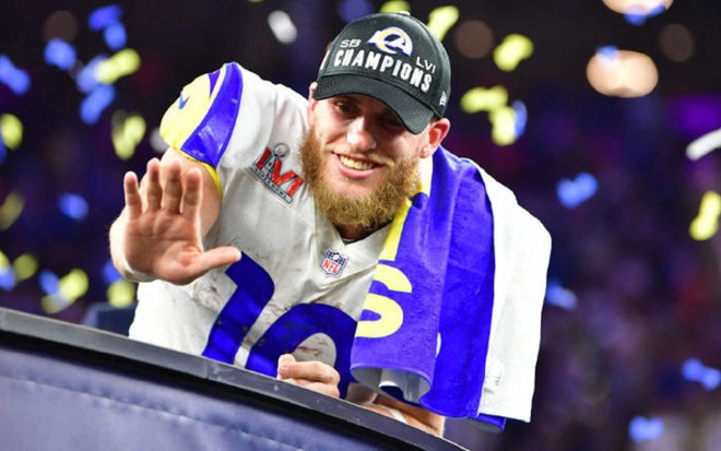 Cooper Kupp com um boné de vitória do SuperBowl com uma camisa branca e acenando para o público