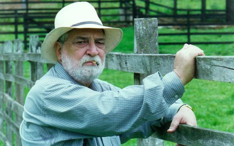 O ator Lafayette Galvão (1931-2019) caracterizado como seu personagem em Corpo Dourado, com camisa azul, chapéu branco, cigarro na boca, com as mãos em uma cerca, num pasto