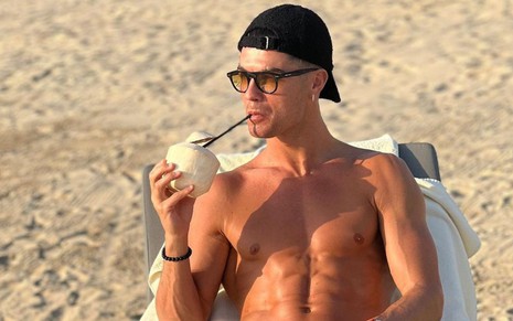Cristiano Ronaldo toma bebida em praia; ele está sem camisa