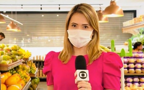 Imagem de Cristina Mayumi em matéria do Jornal Nacional; ela usa blusa rosa, está de máscara e segura microfone com logo da emissora