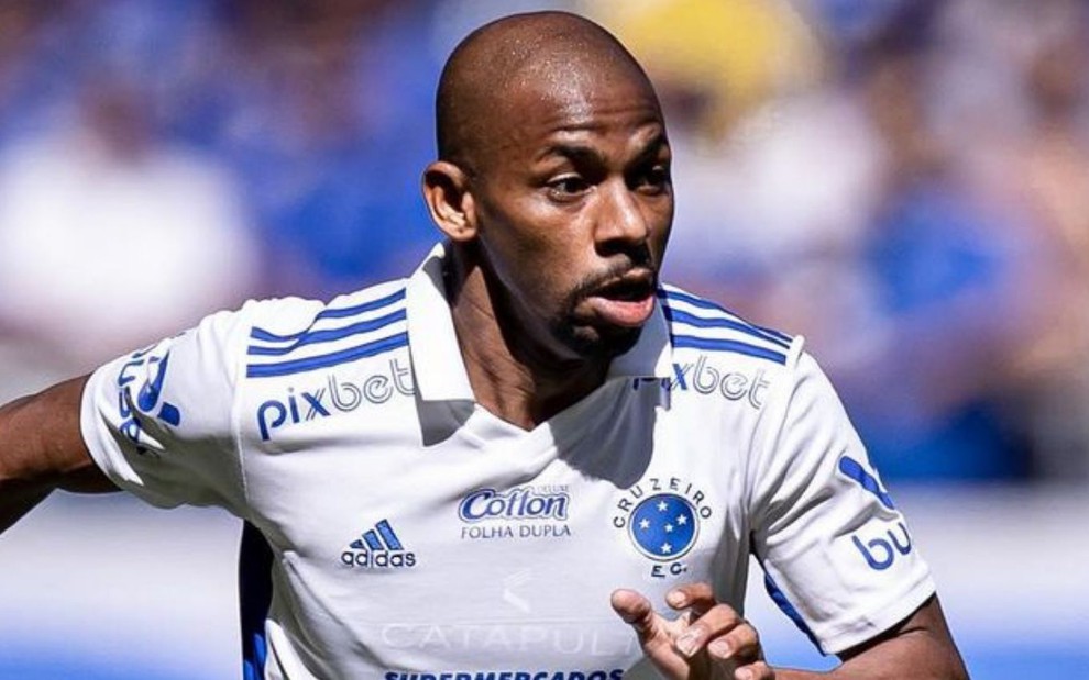 Jogador Waguininho, do Cruzeiro, corre em campo e veste uniforme branco com detalhes azuis