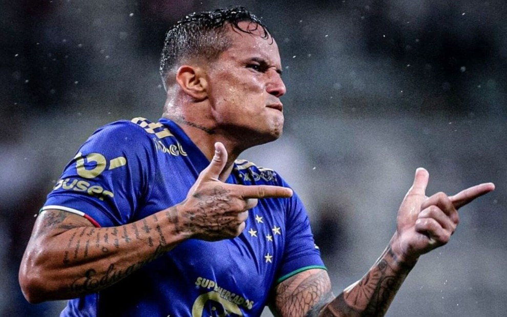 Edu, atacante do Cruzeiro, com a camisa azul do clube mineiro e fazendo um sinal de arma