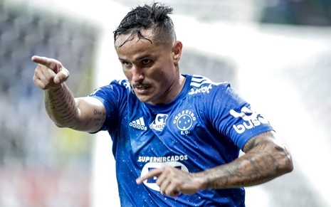 Jogador Edu, do Cruzeiro, aponta para frente e veste uniforme azul com detalhes brancos