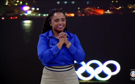 Daiane dos Santos com uma blusa azul, saia marrom e as mãos juntas, nos estúdios da Globo para a transmissão dos Jogos Olímpicos de Tóquio