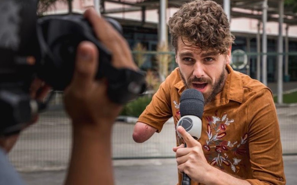 No primeiro plano, uma mão segura uma câmera, o foco da imagem vai no segundo plano, no qual Daniel Toko está vestindo uma camiseta marrom e segura o microfone perto da boca enquanto fala