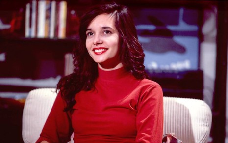 A atriz Daniella Perez de blusa vermelha, sorrindo, em cenário da novela De Corpo e Alma, na Globo