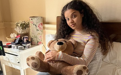 Danielle Olímpia está segurando um ursinho de pelúcia no cenário de seu quarto em Travessia; ela está sentada na cama