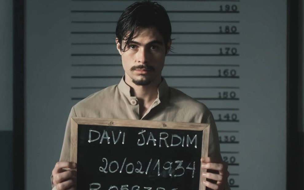 Ator Rafael Vitti segura uma placa com o nome Davi Jardim escrito e olha para a câmera em cena de Além da Ilusão