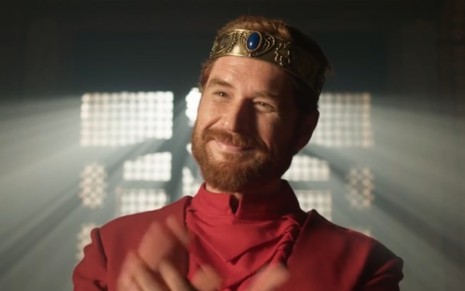 O ator Cirillo Luna sorri em cena da novela Reis, com traje vermelho e coroa de rei