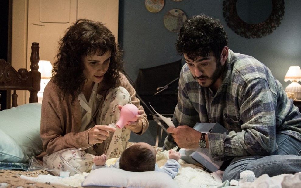 Débora Falabella e Marcos Veras sentados na cama olhando e brincando com um um bebê que está deitado