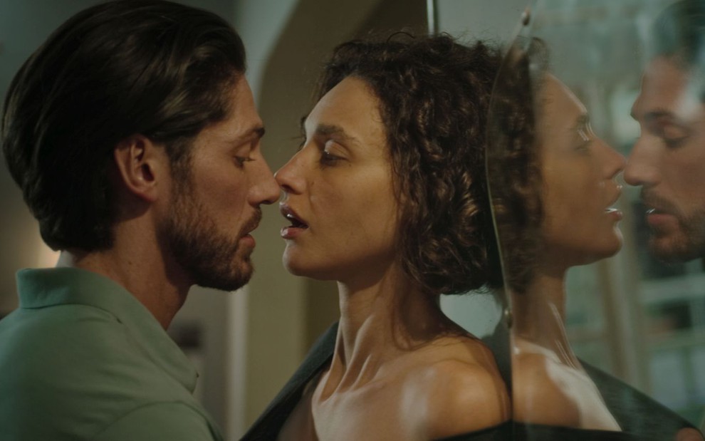 Em cena de Olhar Indiscreto, Débora Nascimento está quase beijando Ângelo Rodrigues; eles estão encostados em um espelho