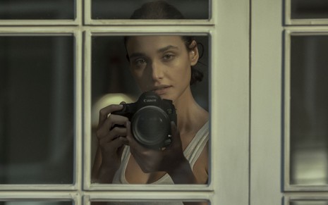 Em cena de Olhar Indiscreto, Débora Nascimento está com uma câmera fotográfica olhando por uma janela