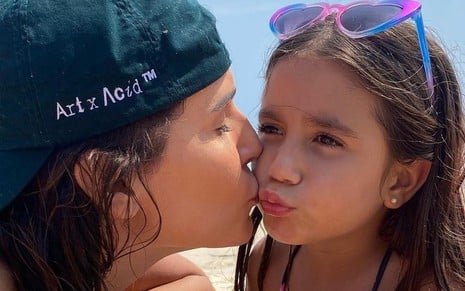 De boné, na praia, Deborah Secco dá um beijo no rosto da filha, Maria Flor, que está com óculos de sol na cabeça, e fazendo biquinho