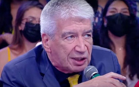 Décio Piccinini como jurado em quadro do Programa do Ratinho, com expressão séria e microfone na mão