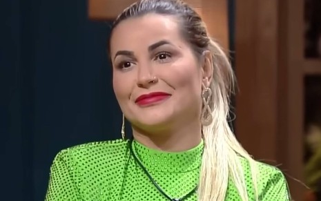 Deolane Bezerra veste roupa verde com gola levemente alta, usa rabo de cabelo e batom vermelho, e faz cara de deboche