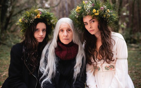 As atrizes Camila Botelho, Cassia Kis e Anna Melo caracterizadas como suas personagens em Desalma, lado a lado, olhando para a câmera, em cenário de floresta, Camila e Anna com coroas de flores