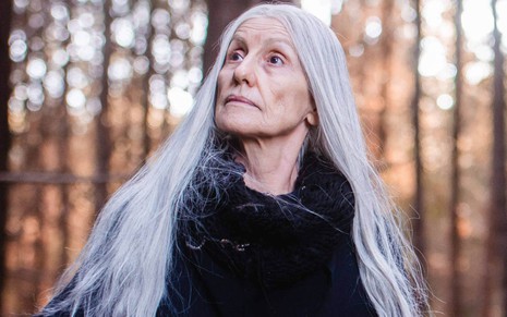 Cassia Kis de cabelos longos brancos, olhando para a esquerda e para cima, em paisagem de floresta, na série Desalma