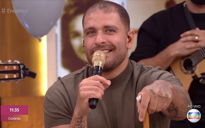Diogo Nogueira no estúdio do Encontro com Fátima Bernardes segurando um microfone na mão esquerda e se apoiando em uma bangala na mão esquerda e um sorriso de canto.