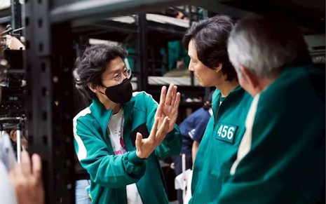 O diretor da série da Netflix Round 6, Hwang Dong-hyuk , está no set conversando com os protagonistas da série. Todos estão de verde, vestindo o uniforme que os personegens vestem em Round 6.