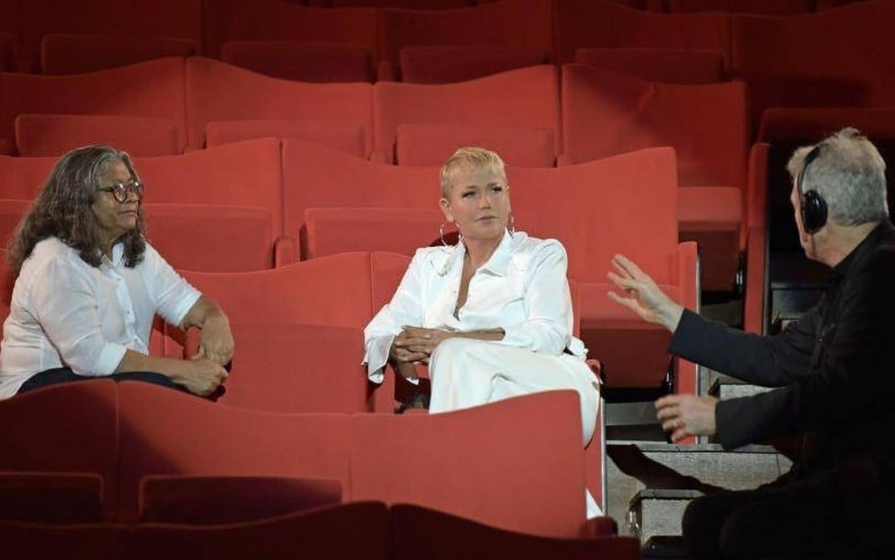 Xuxa Meneghel e Marlene Mattos estão sentadas em um auditório; as duas vestem branco e conversam com um homem de costas