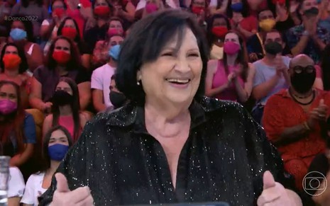Sorridente de roupa preta, Déa Lúcia em frente à plateia do Domingão com Huck