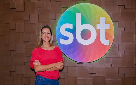 Domitila Becker usa uma camisa rosa com calça azul, está com os braços cruzados e sorri para a câmera ao lado do logotipo do SBT, na sede da emissora em Osasco (SP)