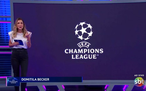 Domitila Becker usa uma camisa branca com calça preta, e está no cenário da Uefa Champions League no SBT onde será a apresentadora das transmissões