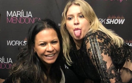 Ruth Moreira e Marília Mendonça posam juntas; a mãe da cantora sorri, enquanto a sertaneja mostra a língua na frente de um banner