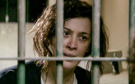 A atriz Claudia Raia como Donatela em A Favorita; ela está descabelada e atrás das grades, olhando com cara de desconfiança