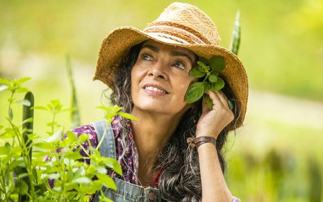Claudia Ohana posa com o figurino de sua personagem em Vai na Fé com chapéu e uma planta na orelha