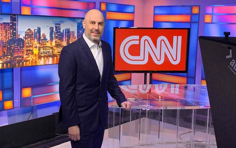 O jornalista Douglas Tavolaro nos estúdios da CNN em Atlanta, nos Estados Unidos; ele veste terno e está ao lado de um grande logotipo da marca