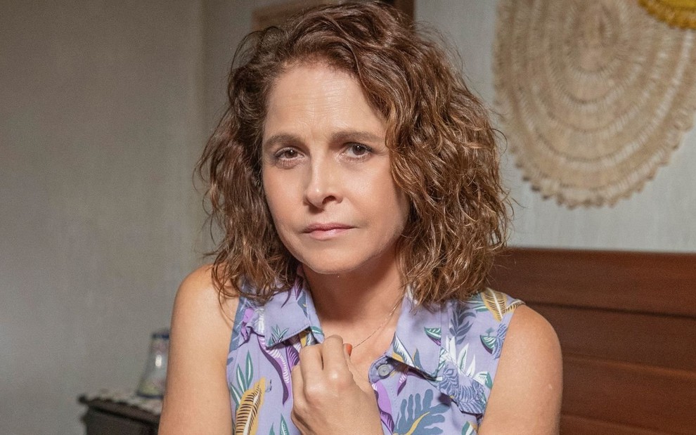 Drica Moraes está caracterizada como sua personagem em Travessia, sentada em um banco