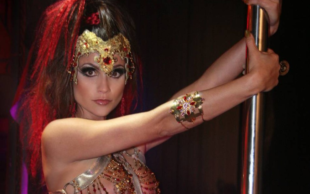 Flávia Alessandra caracterizada como Alzira, com adereço de cabeça, maquiagem forte e segurando em pole dance em cena da novela Duas Caras (2007)