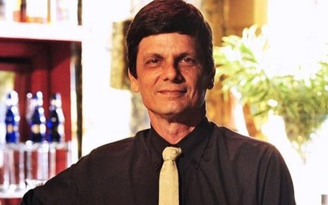 O ator Duda Ribeiro de camisa preta, gravata branca, com leve sorriso de boca fechada em cena de Caminho das Índias