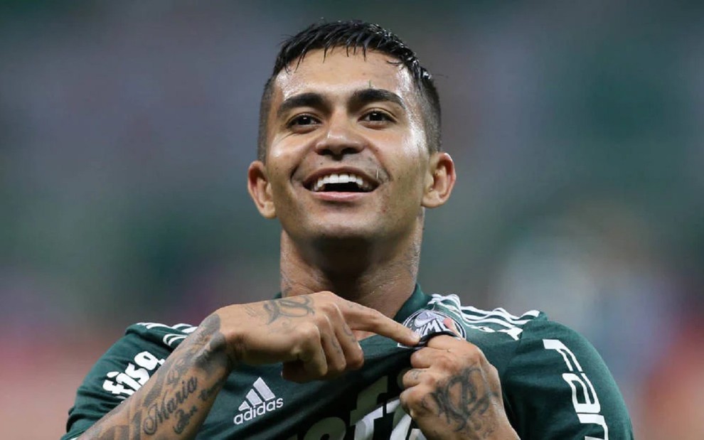 Dudu vibra com gol do Palmeiras. Ele usa uma camisa verde e comemora com seus colegas de banco, que usam um colete amarelo