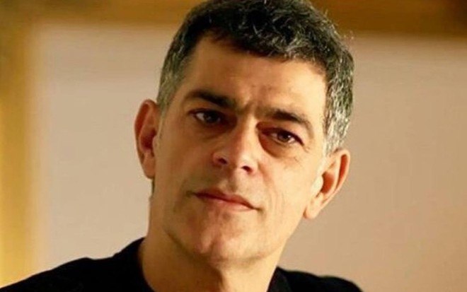 Eduardo Moscovis olha para frente, veste blusa preta e está na frente de uma parede branca