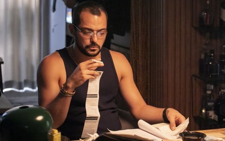 Em cena de Os Outros, Eduardo Sterblitch está usando óculos, fumando e sentado, observando um caderno que está em cima da mesa