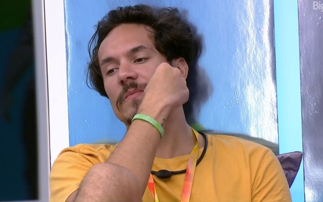 Eliezer Netto no quarto do líder no BBB 22, ele está apoiado na parede, veste uma camiseta marela e exibe um semblante reflexivo