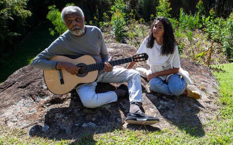 Gilberto Gil e sua filha Bela Gil numa área aberta em meio à natureza, os dois sentados numa pedra, ele segura um violão, durante gravações do programa Em Casa com os Gil