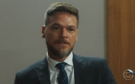Em cena de Vai na Fé, Emilio Dantas está usando terno e gravata e está olhando para alguém