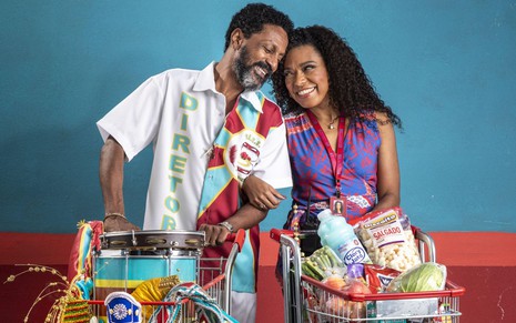 Luis Miranda e Vilma Melo seguram dois carrinhos de supermercado: no dele tem produtos de escola de samba e no dela de mercado