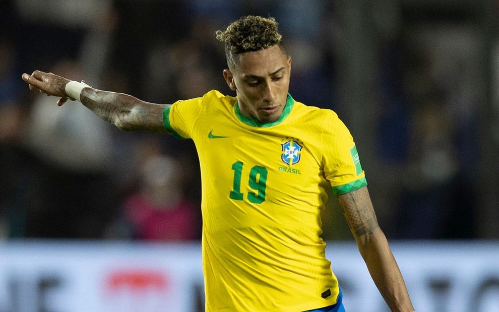 O atacante Raphinha chuta bola em jogo da Seleção Brasileira; ele está com uniforme amarelo e azul