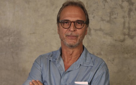 O autor Euclydes Marinho com expressão séria, óculos de grau, camisa azul, posa para foto com fundo bege atrás
