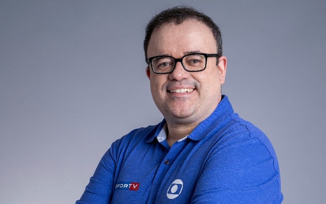 Everaldo Marques, da Globo, com uniforme azul, óculos de grau preto, sorrindo com um fundo cinza