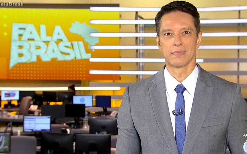 Sergio veste terno cinza, camiseta branca e gravata azul, ele olha para a câmera e está na frente de um fundo amarelo escrito "Fala Brasil"