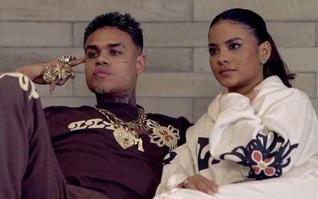 MC Cabelinho usa um conjunto marsala, e Bella Campos veste um moletom branco; eles estão sentados no sofá de casa, sérios, durante entrevista ao Fantástico